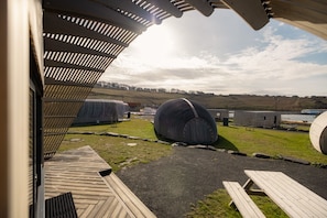 Vista desde un espacio exterior cubierto con una terraza de madera, una estructura cilíndrica grande y un edificio con un diseño de techo particular.