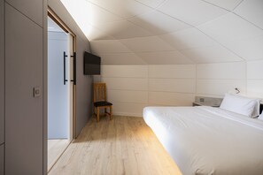 Un dormitorio con cama, una silla, televisor montado en la pared y una puerta que da a otra habitación.