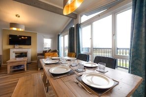 A dining area with a wooden table set for four, a television mounted on the wall, and a view of the outdoors through large windows. - Lost Shore Surf Resort (Newbridge)