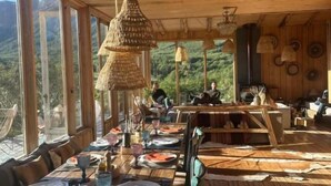 Un comedor de madera con mesas dispuestas para una comida, ventanas amplias y vistas al exterior.