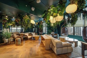 Eine moderne Lounge mit Holzböden, bequemen Sitzgelegenheiten, hängenden Pflanzen und großen Fenstern mit Blick auf die Stadt.