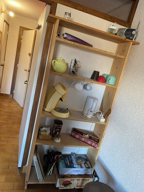 Ein Holzbücherregal mit Küchenutensilien, einer Teekanne, einem Wasserkocher, einer Kaffeemaschine und einigen Büchern.