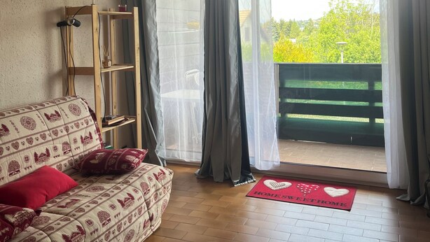 Ett vardagsrum med en mönstrad soffa, en hylla i trä, en rund, vävd matta och utsikt över ett grönområde utomhus genom en skjutdörr.