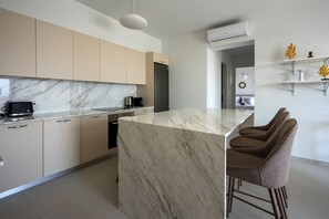 A modern kitchen with marble countertops, a built-in oven, and a bar area with stools. - Aurea Kalithea Sunset  Sunrise  Sea (Asomatos)