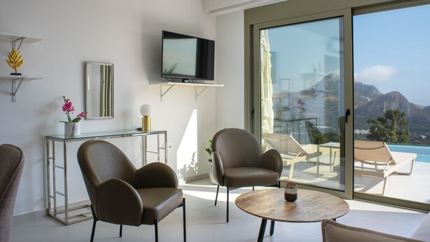 Un soggiorno moderno con televisore piatto, due poltrone, un tavolino rotondo e un'ampia porta scorrevole in vetro che dà su un balcone con vista mare.