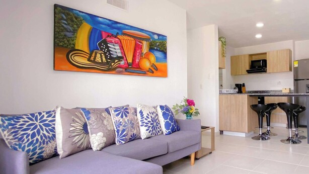Ein Wohnzimmer mit einer Couch, bunten Kissen, einem Gemälde und einer Küche im Hintergrund.
