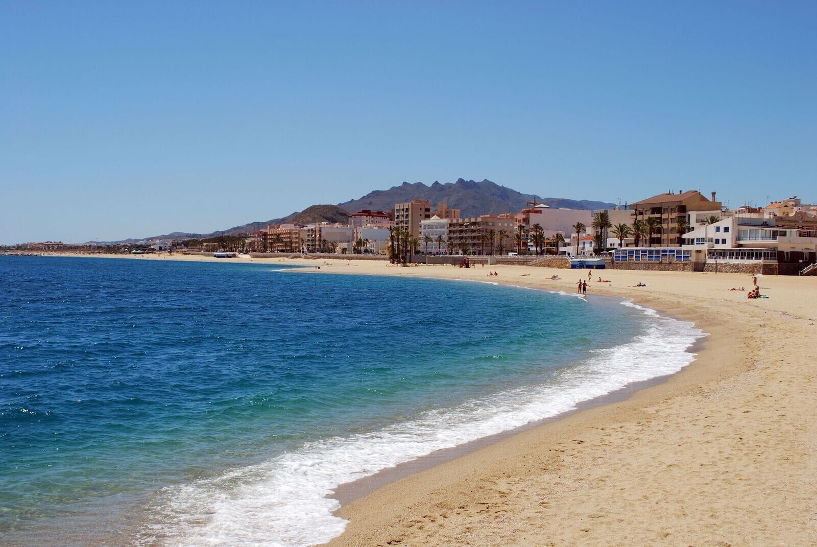 A beach with clear blue water, a sandy shore, and buildings in the background.
