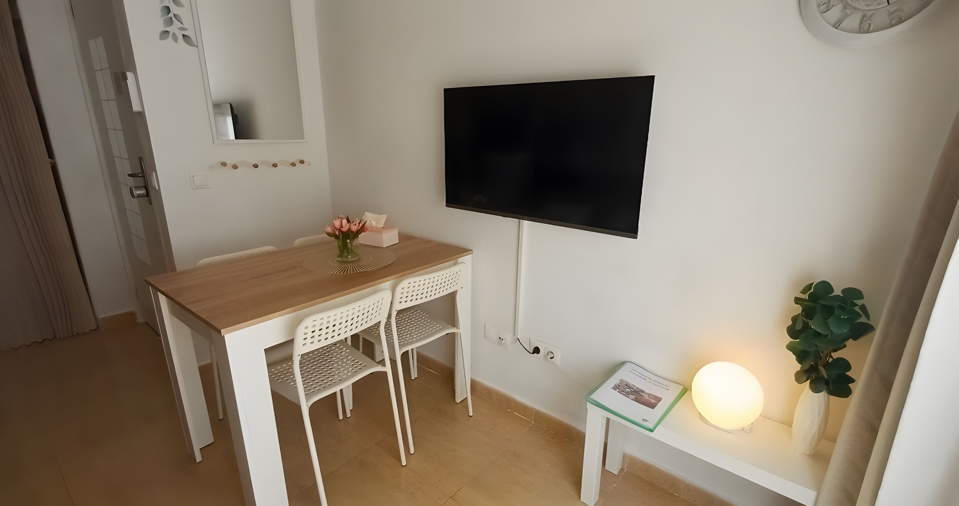 Een kamer met een houten bureau, twee witte stoelen, een flatscreen televisie, een klein tafeltje met een lamp en een potplant.