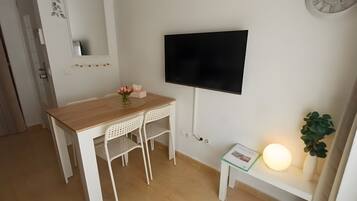 Een kamer met een houten bureau, twee witte stoelen, een flatscreen televisie, een klein tafeltje met een lamp en een potplant.