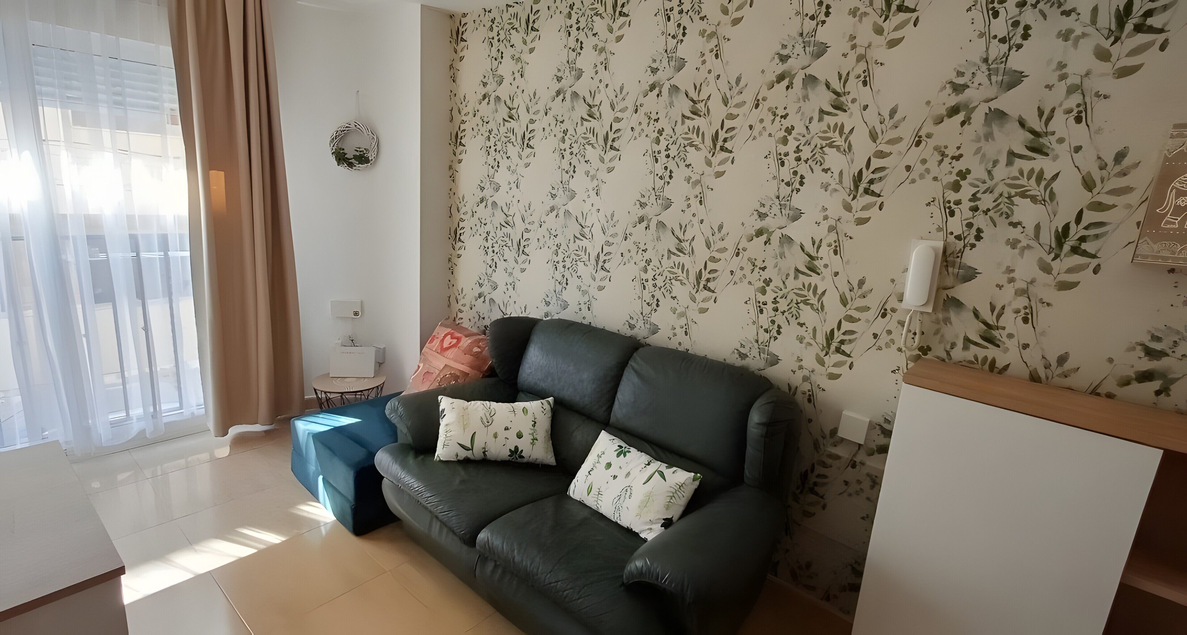 Una sala de estar con un sofá de cuero oscuro, dos cojines a juego, una mesita redonda con una caja y un teléfono fijado a la pared.
