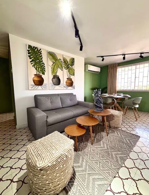 A living room with a grey sofa, two round wooden tables, a patterned rug, and a wall-mounted air conditioner.