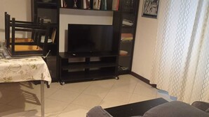A living room with a television, a dining table with chairs, a sofa, a bookshelf, and a patterned curtain.