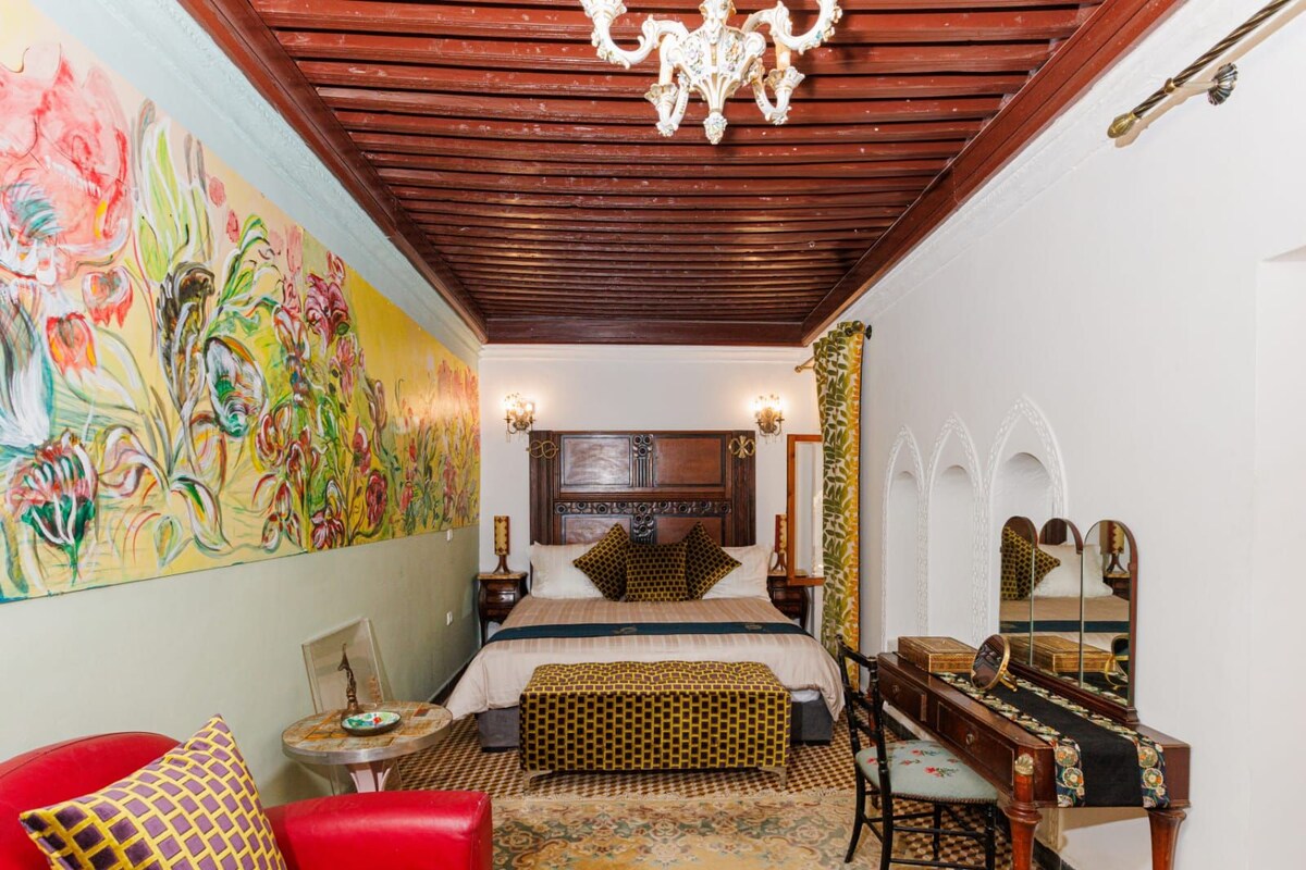 Um quarto com teto de madeira, cama com roupa de cama estampada, sofá vermelho, escrivaninha com espelho e um painel de parede com pintura floral colorida.