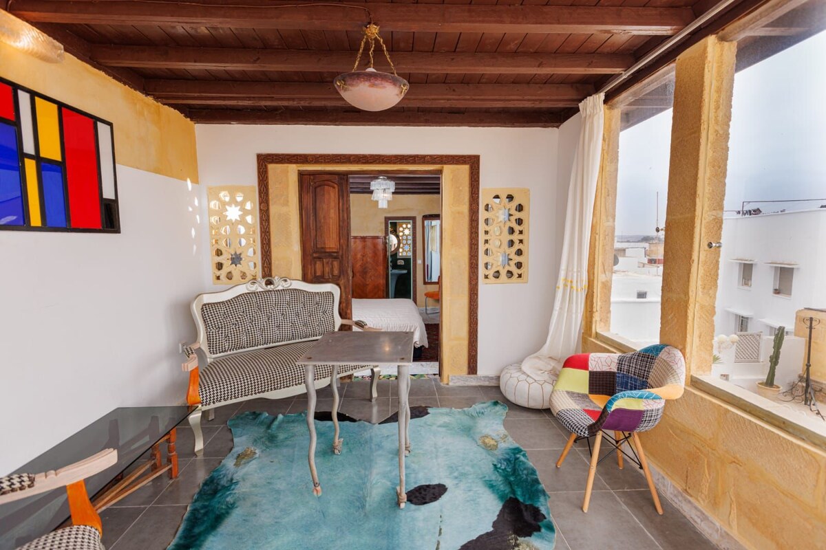 Quarto com teto de madeira, sofá, mesa e vista para edifícios.