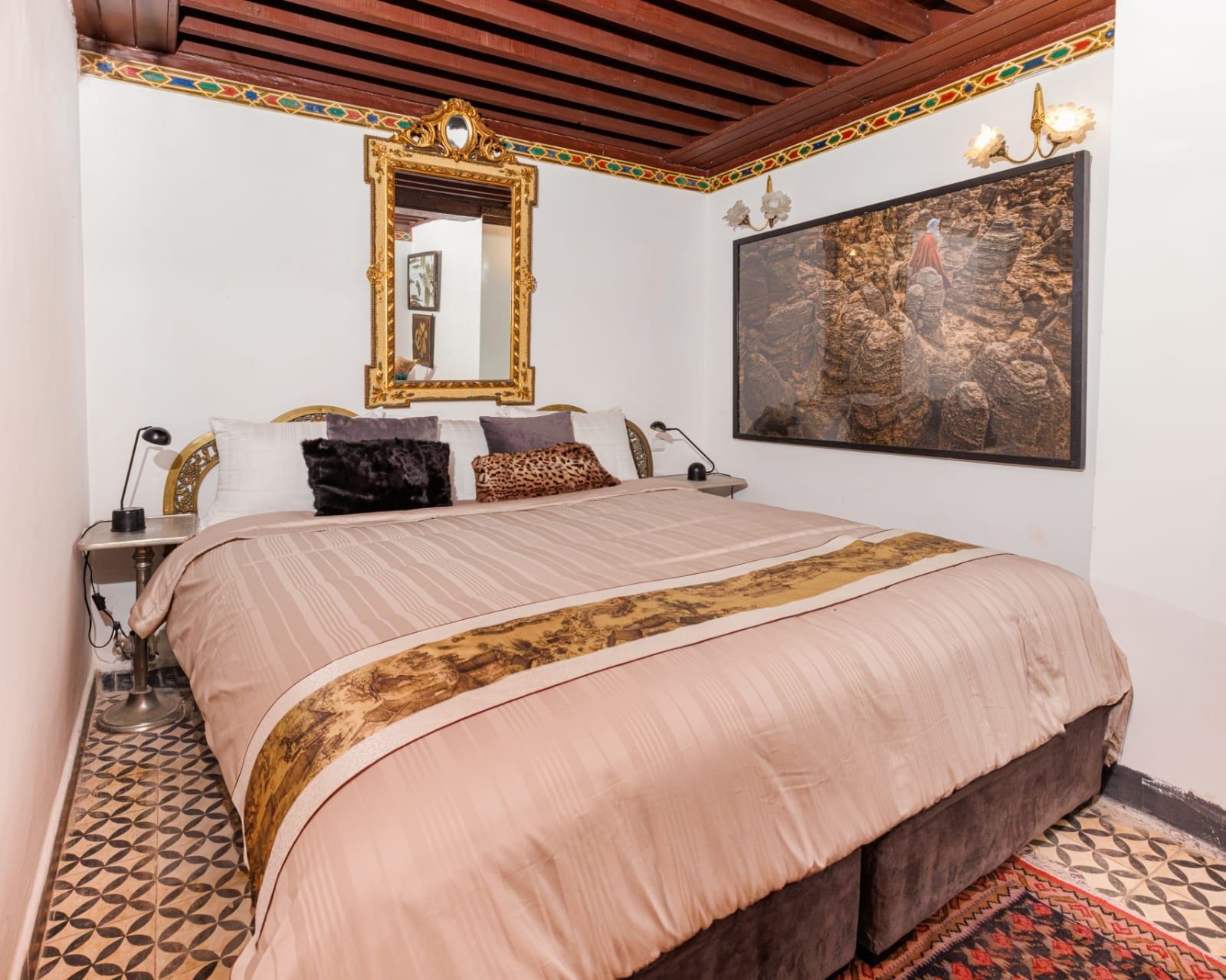 A bedroom with a large bed, a mirror, a wall-mounted artwork, and a patterned rug.