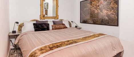 A bedroom with a large bed, a mirror, a wall-mounted artwork, and a patterned rug.
