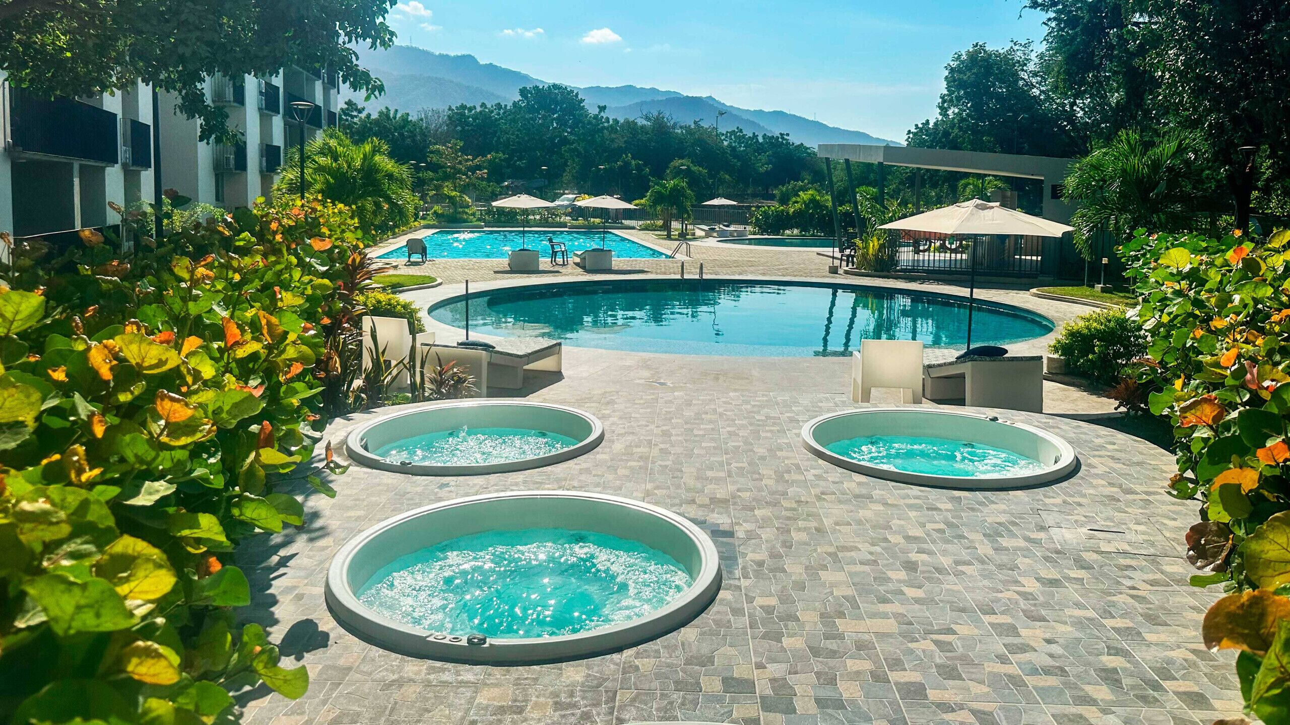 A pool area with a large central pool and three smaller hot tubs, surrounded by greenery and a mountainous backdrop.