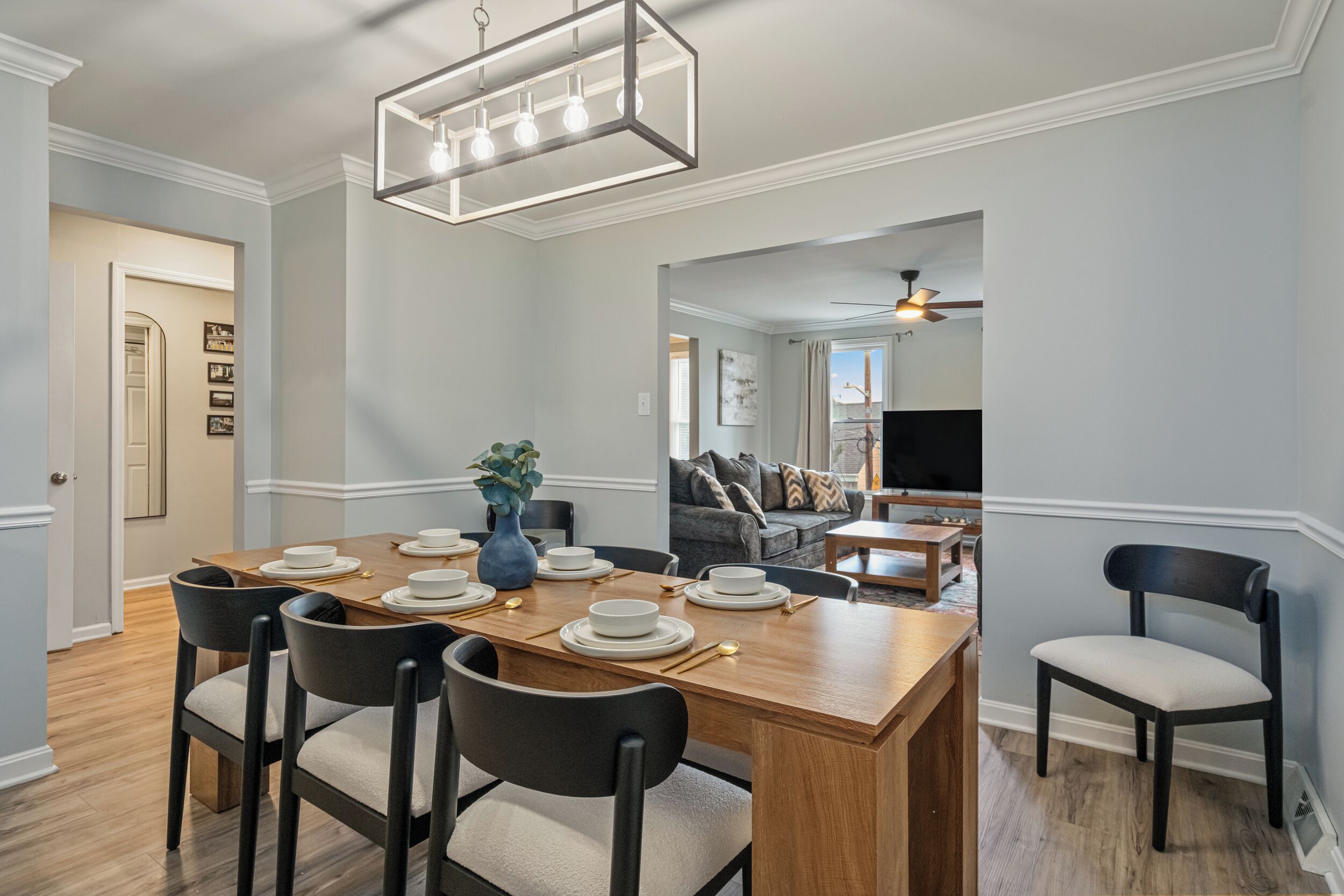 A dining area with a wooden table set for six, surrounded by chairs, a vase with greenery, and a modern light fixture above.