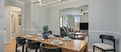 A dining area with a wooden table set for six, surrounded by chairs, a vase with greenery, and a modern light fixture above.