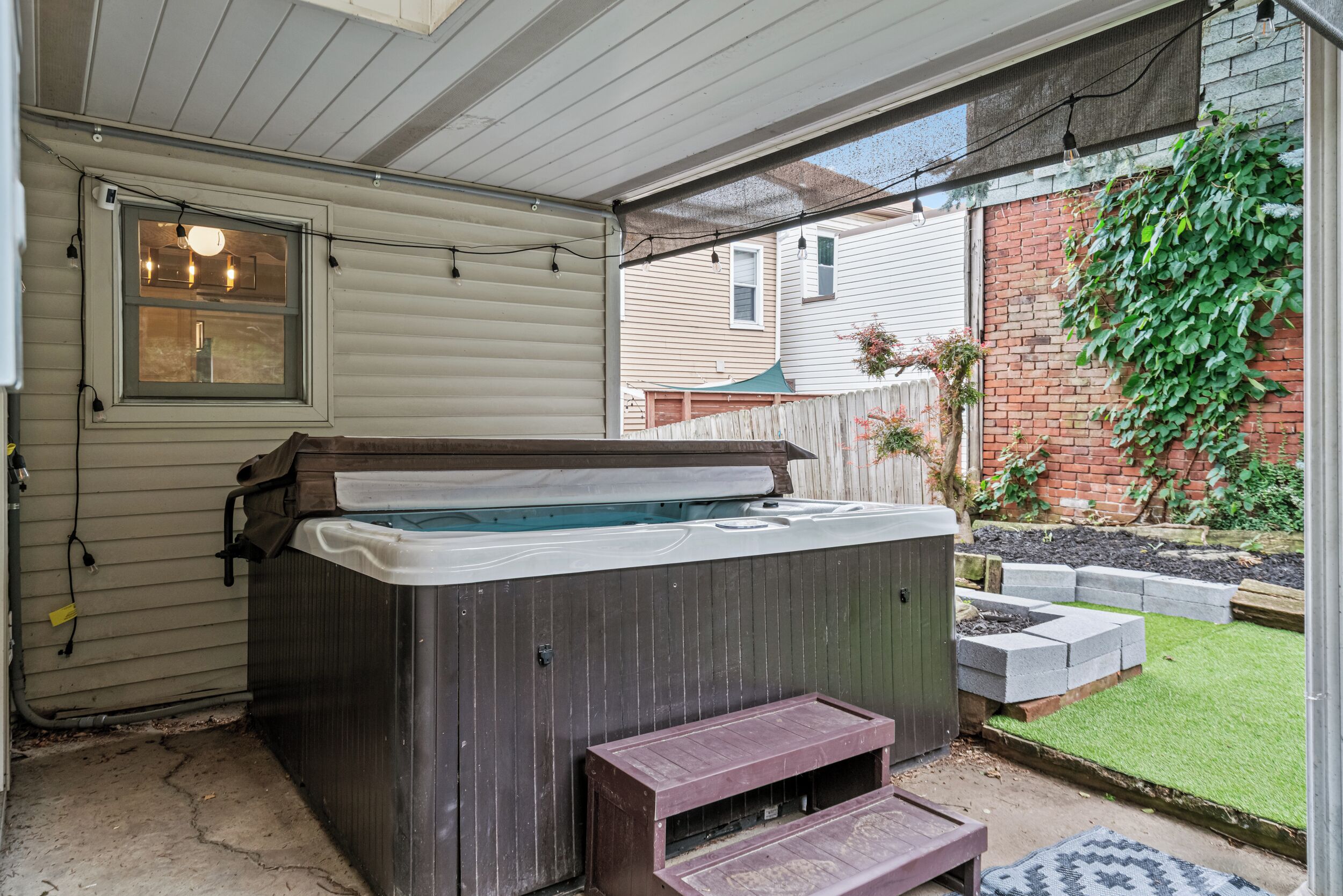 Un bain à remous situé dans un espace extérieur abrité, doté d’une terrasse en bois, d’une fenêtre et d’un mur en briques agrémenté de plantes.