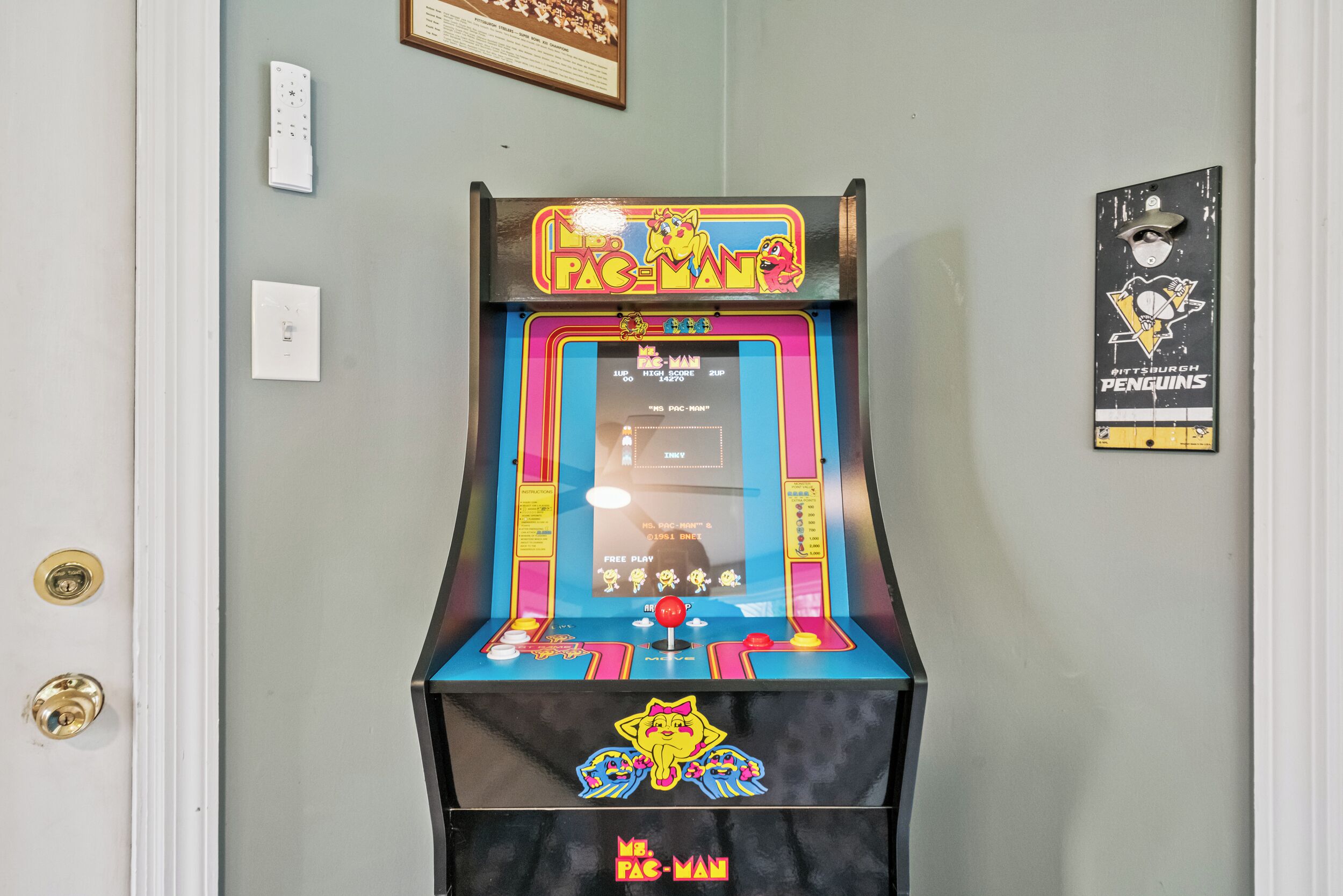En gammal Pac-Man-spelautomat i ett rum med ljusgröna väggar, en inramad tavla och en dörr med ett handtag av mässing.