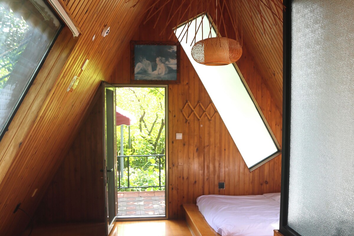 A room with a sloped ceiling, a bed, a small table, a bench, and a view through a window.