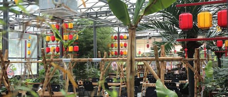 A greenhouse-style restaurant with bamboo structures, tables, chairs, and red lanterns.