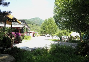 A parking area with a building on the left, surrounded by greenery and a mountain in the background.