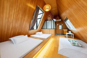 Um quarto acolhedor em formato triangular, com paredes de madeira, duas camas, uma mesa pequena e uma janela.