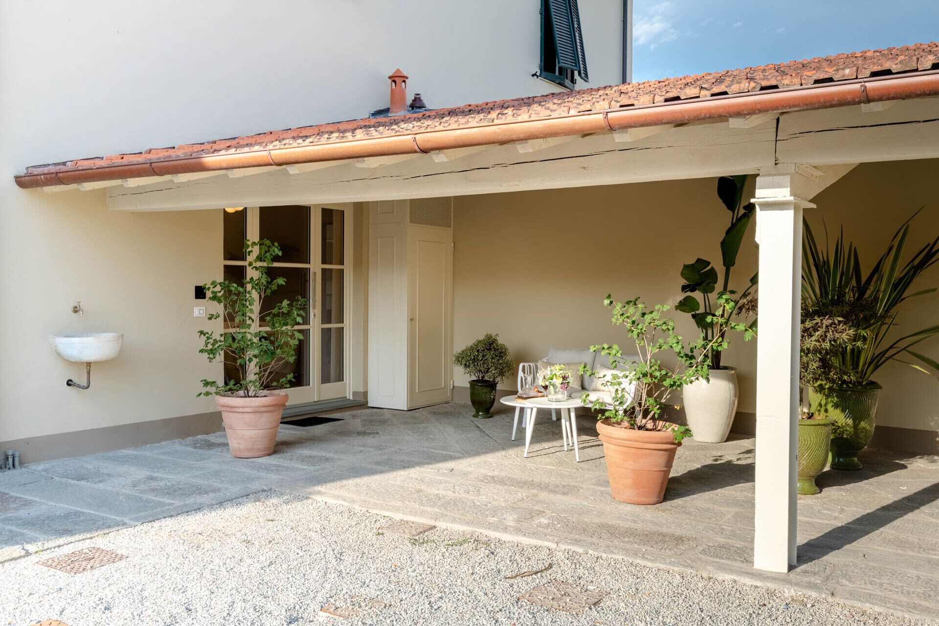 A covered outdoor area with a tiled roof, potted plants, a small table, and a white sink against the wall.