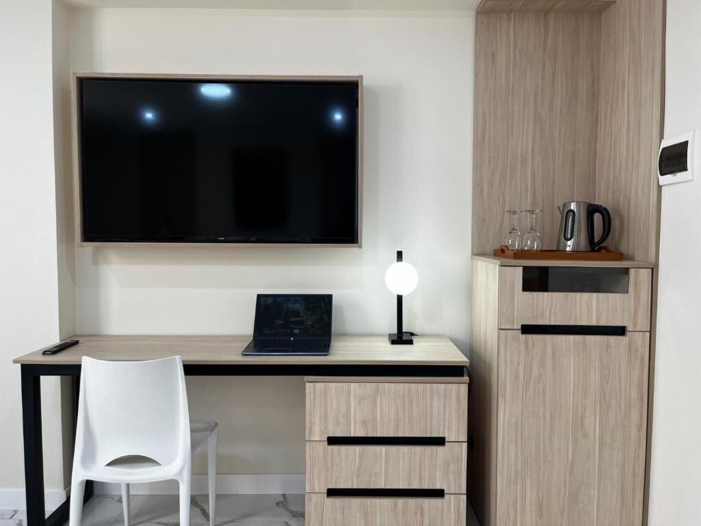 A modern room with a flat-screen TV, a desk with a laptop, a white chair, and a wooden cabinet with a kettle and glassware.