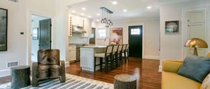 A spacious living area with a dining table, a sofa, and a chair. There is a kitchen in the background with modern appliances and a dining island.