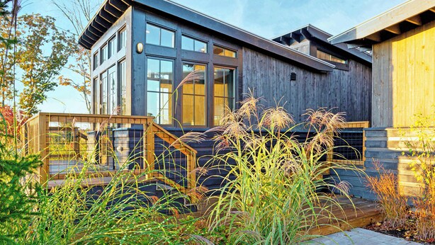 Une maison moderne à deux étages, dotée de grandes fenêtres, d’un revêtement extérieur en bois et d’une terrasse en bois.
