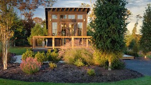 Une maison moderne à deux étages, dotée d’un balcon, et entourée d’un jardin aménagé avec différents arbustes et arbres.