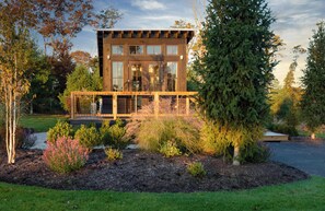 A modern two-story house with a balcony, surrounded by a landscaped garden with various shrubs and trees.