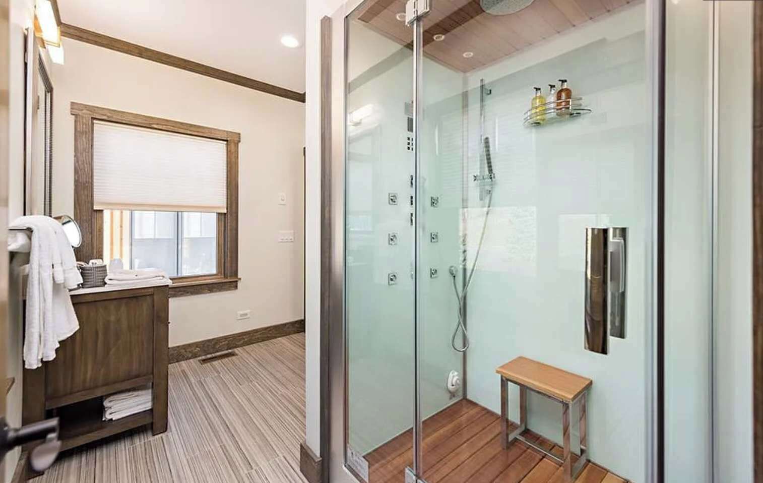 Un baño moderno con cabina de ducha de vidrio, un banco de madera y un tocador con espejo y toallero.