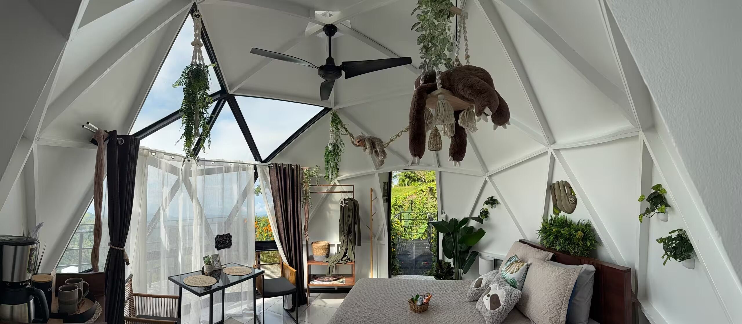 A geodesic dome room with a bed, a table, and hanging plants.