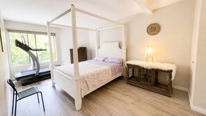 Una cama con dosel y piecera, una mesita de noche con lámpara, una silla y una ventana con vistas a la vegetación.
