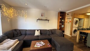 Una sala de estar con un sofá modular, una mesa de centro con revistas, un estante montado en la pared con objetos decorativos, y un área de lavandería con lavadora.
