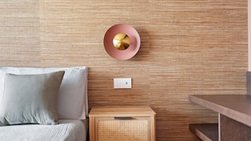 A room with a textured wall, a round wall-mounted light fixture, a bedside table with a drawer, and a bed with pillows.