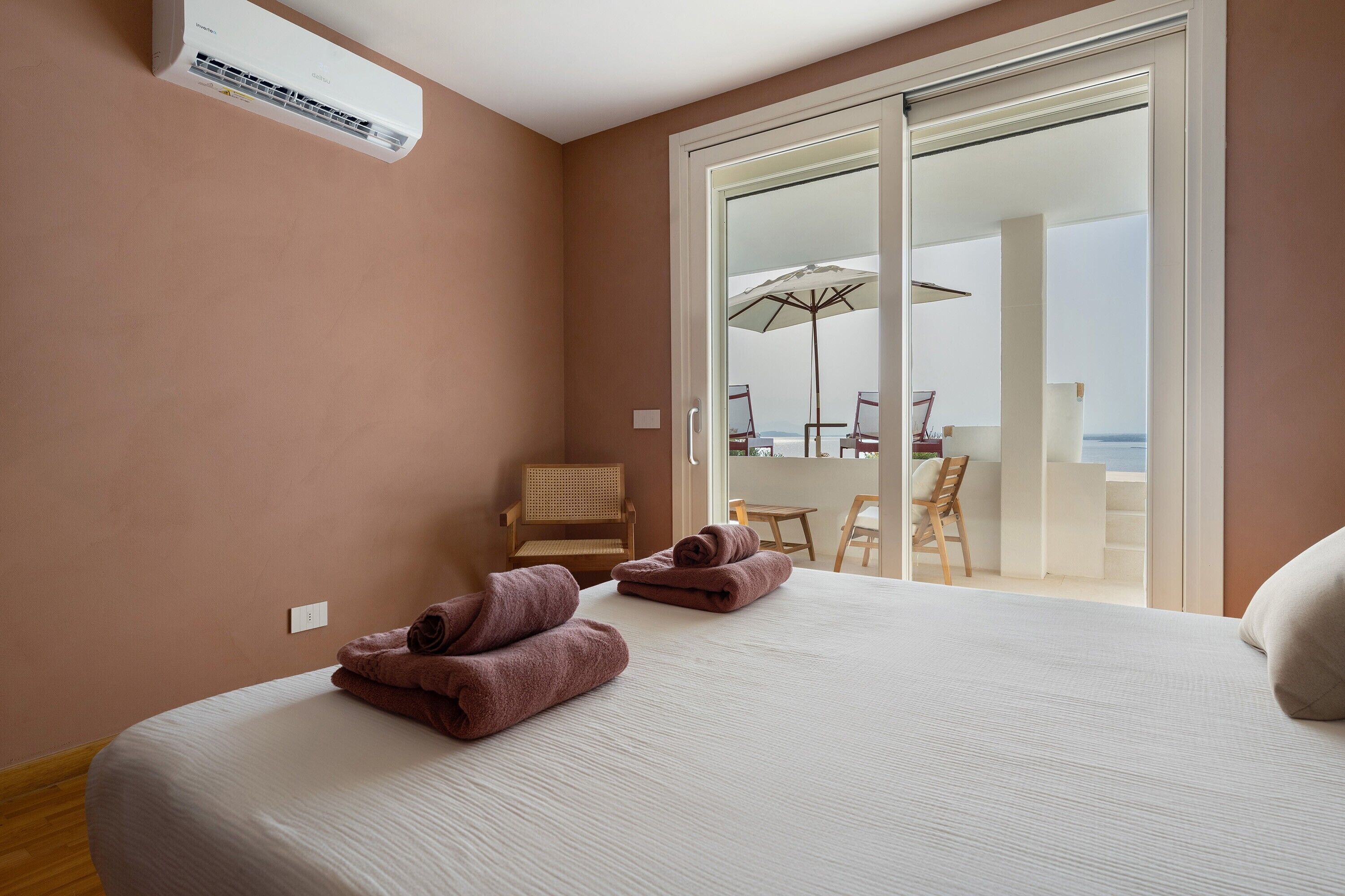 A bedroom with a bed, a chair, and a view of a balcony with outdoor furniture.