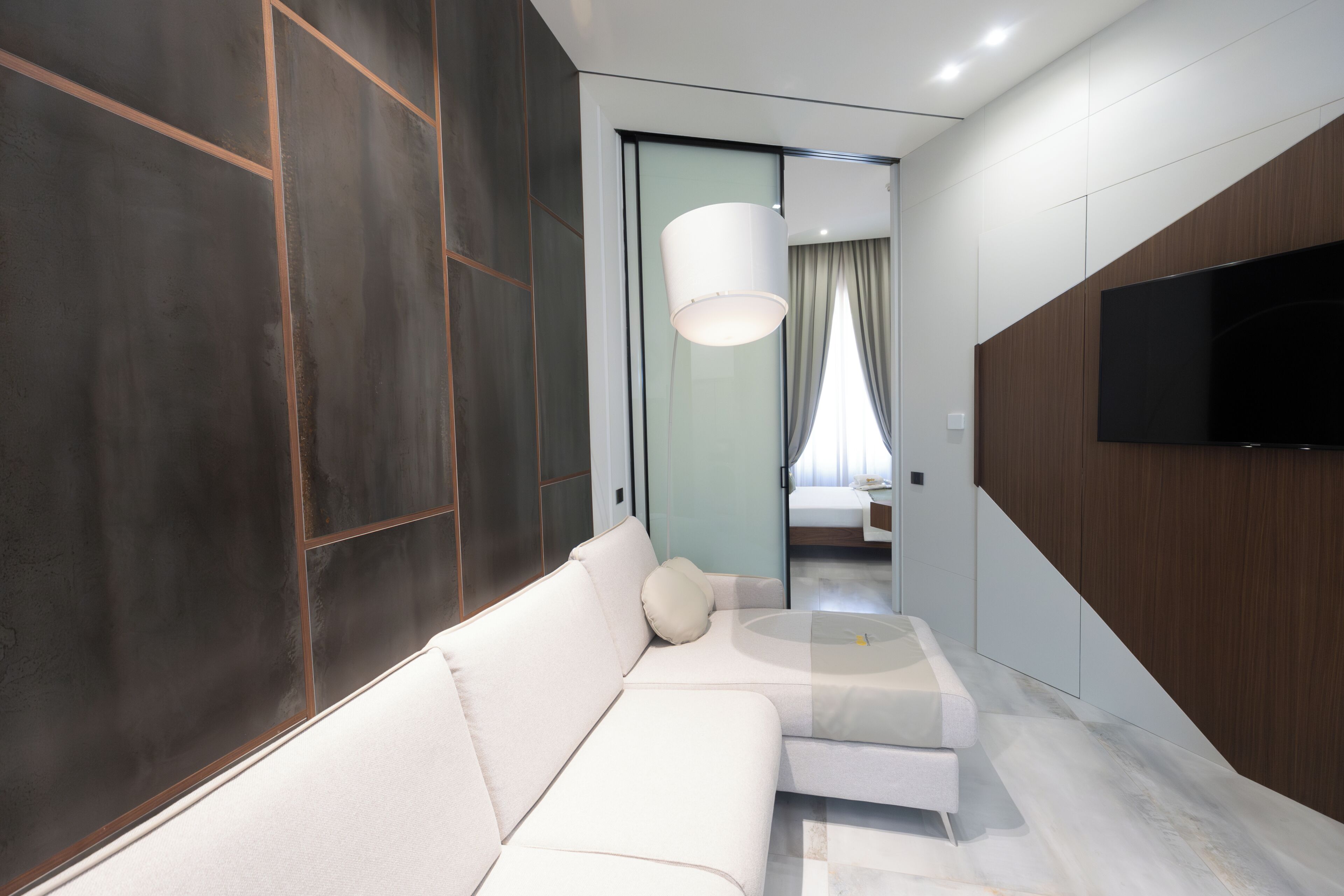 Una moderna camera d'albergo con un divano bianco, una TV a schermo piatto montata a parete e un'ampia finestra con tende.