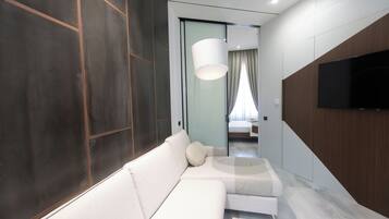 Quarto de hotel moderno com sofá branco, televisão de tela plana na parede e uma janela grande com cortinas.
