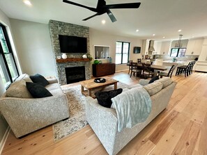 A spacious living room with a fireplace, a large sofa set, a dining area, and a kitchen in the background. - Bolton Creekside Condo - Walk to lake, beaches, marinas, restaurants, shops (Bolton Landing)