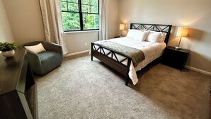 Ein Schlafzimmer mit einem Bett, einem Sessel, einem Nachttisch, einer Lampe und einem Fenster mit Vorhängen.