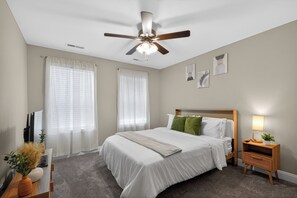 A bedroom with a bed, a ceiling fan, a bedside table with a lamp, and a wall-mounted artwork.