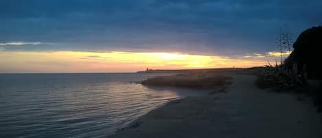 Una playa con un atardecer, mar tranquilo y un promontorio rocoso.