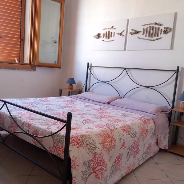 Ein Schlafzimmer mit einem Bett, einem Nachttisch, einer Lampe, einem Fenster mit Jalousien und Wandbildern.