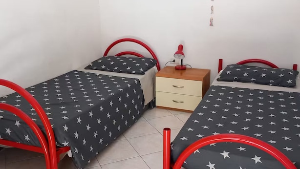 Een kamer met twee eenpersoonsbedden met rode metalen frames, een klein houten nachtkastje en een rode bureaulamp.