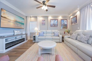 Een woonkamer met een plafondventilator, een grote flatscreen-tv, een bank, een salontafel en een kamerkleed met dessin.